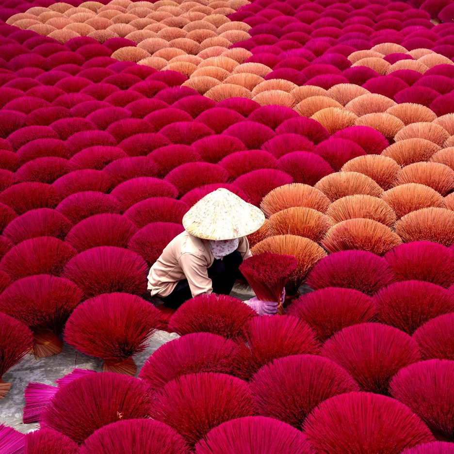 Craftsperson in a conical hat among vibrant magenta and peach incense bundles, Vietnam
